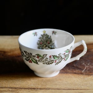 May include: A white ceramic teacup featuring a green holly and red berry garland pattern around the exterior. The interior of the cup displays a detailed illustration of a decorated Christmas tree with a yellow star topper and small holly accents.