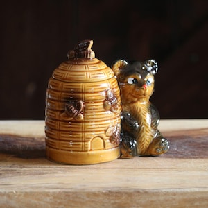 May include: A ceramic honey pot shaped like a beehive with a matching bear figurine. The honey pot is tan with brown accents and bee details. The bear is brown and black with a friendly expression. The items are on a wooden surface.