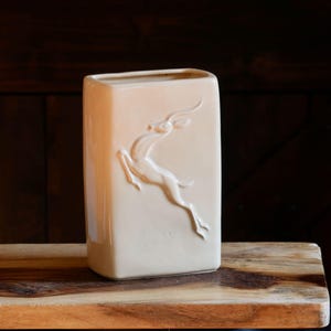 May include: A rectangular, cream-colored vase with a raised relief of a leaping gazelle. The vase is displayed on a wooden surface, with a dark brown background. The vase is approximately 8 inches tall.