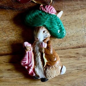 May include: A ceramic figurine of a rabbit wearing a green hat and brown coat, holding a pink bag. The rabbit has a white face and ears, with black stripes. The figurine is on a wooden surface.
