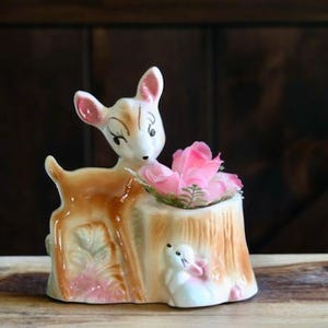 May include: A ceramic deer figurine with a fawn-like appearance, featuring a pink-colored flower arrangement in a tree stump planter. The figurine has a white and brown color scheme with pink accents. A small white rabbit is at the base.