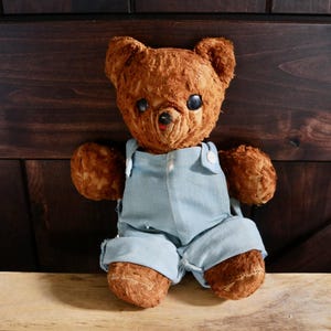 May include: A brown teddy bear with textured fur, black plastic eyes, and a small stitched nose. The bear is wearing light blue denim-style overalls with white buttons on the straps. The toy has a rounded head, small ears, and articulated limbs.