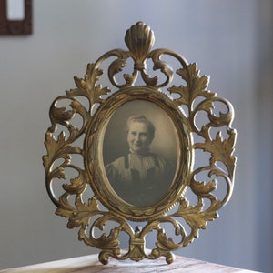 May include: An antique gold-colored oval picture frame with an ornate, leaf-like design. The frame holds a black and white portrait of a woman. The frame is displayed on a marble surface.