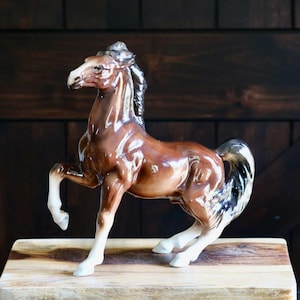 May include: A glossy brown ceramic horse figurine with a raised front leg. The horse has a white mane and tail, and is posed on a wooden block. The figurine is a decorative item.