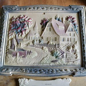 May include: A framed, three-dimensional relief carving depicting a village scene with houses, trees, and a winding road. The artwork is in muted tones of beige, brown, and green, with a decorative border. The frame is approximately 12 inches wide.