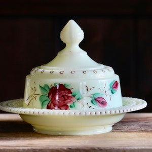 May include: A pale yellow, covered butter dish with a floral design. The dish features a base, a domed lid, and a decorative finial. The floral design includes red roses and pink buds with green leaves.