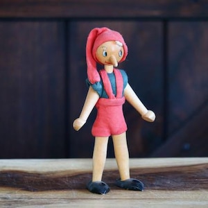 May include: A wooden Pinocchio doll stands on a wooden surface. The doll has a long nose, a red outfit, and a matching hat. The doll's arms and legs are made of wood, and it has black shoes. The background is a dark wooden wall.