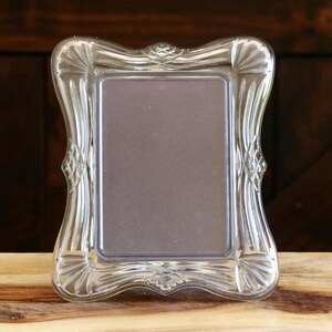 May include: A clear, rectangular picture frame with an ornate, curved design. The frame is made of glass or a similar transparent material and sits on a wooden surface. The frame's design includes decorative flourishes.