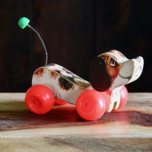 May include: A vintage pull-along toy dog with a spotted body, brown ears, and a green antenna. The dog has red wheels and is made of wood and plastic. A classic toy.
