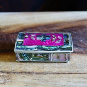 May include: A rectangular jewelry box with a silver frame and a lid decorated with a vibrant pink center and iridescent abalone shell accents. The box sits on a wooden surface.