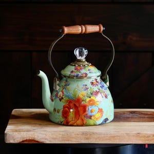 May include: A light green enamel tea kettle featuring a vibrant floral pattern with red, orange, yellow, and blue flowers. The kettle has a curved spout, a clear glass knob on the lid, and a metal handle with a smooth, light brown wooden grip.