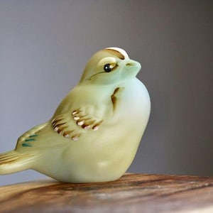 May include: A ceramic bird figurine in shades of pale yellow and white, with brown and green accents. The bird is perched on a wooden surface, with a neutral background. The figurine has detailed wings and a small beak, showcasing a delicate design.