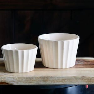 May include: Two off-white ceramic bowls with vertical ridged detailing sit on a wooden cutting board. The bowls are different sizes, with the larger one on the right. The background is a dark wooden surface.