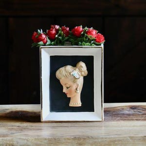 May include: A vintage rectangular vase with a white frame and gold trim, featuring a profile of a woman's face. The vase is filled with red roses. The background is dark wood.