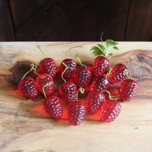 May include: A collection of red glass raspberry ornaments with green stems and leaves, arranged on a wooden cutting board. The ornaments have a textured surface and are illuminated, creating a vibrant display.