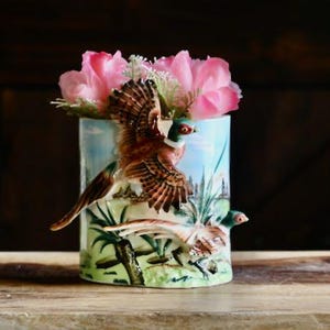 May include: A cylindrical ceramic vase with a pheasant design. The vase features a brown and white pheasant in flight, with a second pheasant on the base. Pink flowers are arranged inside the vase. The background is a light blue with green foliage.