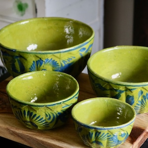 May include: Four handmade ceramic bowls in varying sizes, each with a vibrant lime green glaze and intricate blue floral patterns. The bowls are arranged on a wooden surface, showcasing their unique design and craftsmanship.