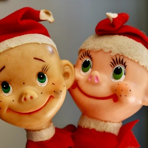May include: Two vintage Christmas elf figurines. Each elf has a smiling face with green eyes, freckles, and a red and white hat. They are wearing red outfits. The figurines are close together.