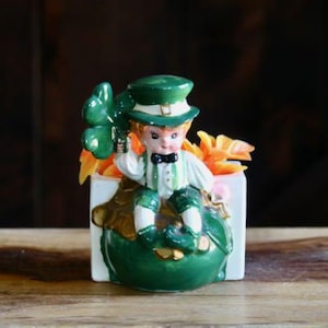May include: A ceramic figurine of a leprechaun holding a shamrock. The leprechaun is wearing a green hat, coat, and pants, with a white shirt and black bow tie. The figurine is set in a white planter with orange and yellow leaves.