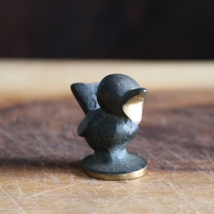 May include: A small, dark-colored bird figurine with a gold-colored beak and base. The bird is perched on a round base, and the background is a blurred wooden surface. The figurine has a vintage or antique appearance.