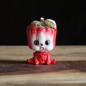 May include: A vintage ceramic figurine of a red pepper with a white face, blue eyes, and a surprised expression. The pepper has green leaves on top and is wearing a red outfit. The figurine is sitting on a wooden surface.