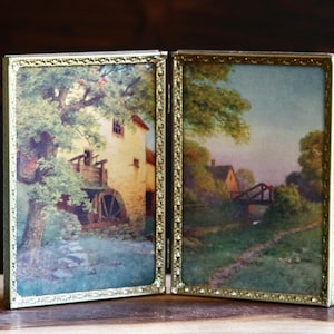 May include: A hinged double picture frame with a gold-toned metal border featuring an embossed pattern. The left panel displays a painting of a water mill and trees, while the right panel shows a landscape painting of a house, bridge, and path.