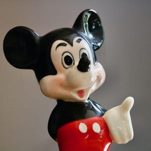 May include: A ceramic Mickey Mouse figurine. The figure has a black head and ears, a white face with rosy cheeks, and a red shorts. Mickey is giving a thumbs up with his white gloved hand.