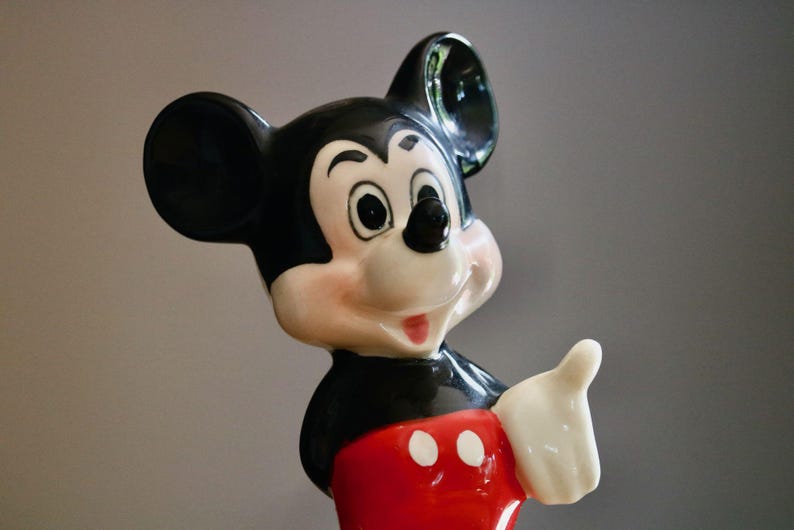 May include: A ceramic Mickey Mouse figurine. The figure has a black head and ears, a white face with rosy cheeks, and a red shorts. Mickey is giving a thumbs up with his white gloved hand.