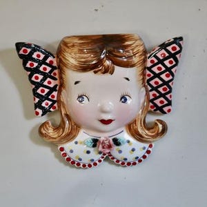 May include: A vintage ceramic wall pocket in the shape of a girl's face. The girl has blonde hair, blue eyes, and red lips. Her collar is white with blue polka dots and a pink rose. The wings are black with red and white diamond patterns.
