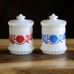 May include: Two white, lidded jars with floral designs. One jar has a red floral pattern, and the other has a blue floral pattern. The jars are on a wooden surface, with a dark background.