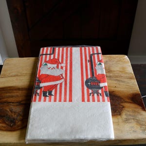 May include: A stack of Christmas-themed paper napkins. The napkins feature a red and white striped pattern with illustrations of Santa Claus and a vintage stove. The bottom portion of the napkins is white.