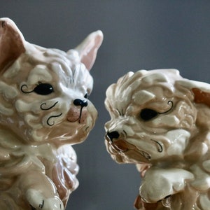 May include: Two ceramic dog figurines in a light beige color with detailed facial features. The dogs have black accents around their eyes and on their snouts. The figurines are facing each other, creating a charming display.