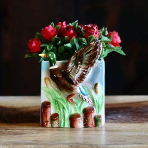 May include: A rectangular ceramic vase with a duck design, filled with red roses. The vase features a duck in flight over a marsh scene. The duck has brown and white wings, a green head, and a blue accent.