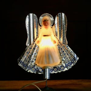 May include: A vintage Christmas tree topper in the shape of an angel. The angel has a white dress, a gold belt, and a crown. The wings are silver with blue and white accents. The angel is lit from within, creating a warm glow.