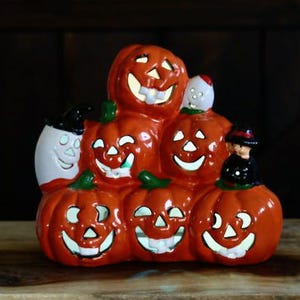 May include: A ceramic Halloween decoration featuring several orange pumpkins with carved smiling faces. A ghost and a figure in a witch hat are also included. The pumpkins are stacked together, creating a festive and spooky display.