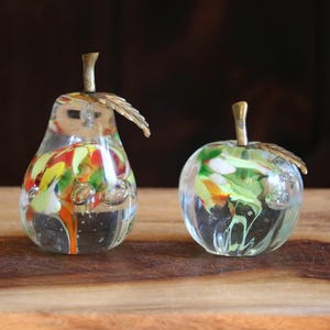 May include: Two clear glass fruit figurines, a pear and an apple, featuring internal swirls of red, yellow, and green colors with embedded air bubbles. Each piece is topped with a textured brass stem and a single brass leaf attached to the top.
