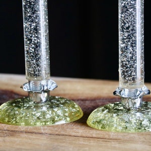 May include: Two clear, cylindrical stems filled with silver glitter particles, each attached to a circular, pale yellow base embedded with silver flakes. The stems feature a silver-toned, ruffled metal collar at the point of connection to the base.