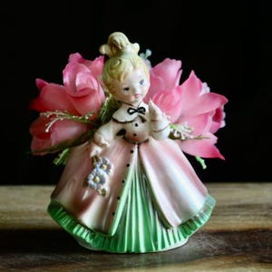 May include: A vintage porcelain figurine of a young girl in a pink and green dress, holding a bouquet of blue flowers. The figurine is surrounded by pink flowers, creating a charming and decorative piece.