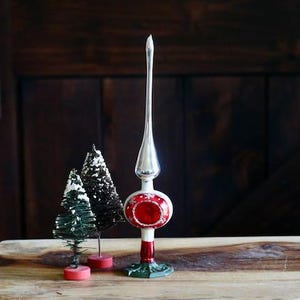 May include: A vintage Christmas tree topper with a silver spire and a red and white globe. Two small decorative Christmas trees are also present. The items are displayed on a wooden surface against a dark wood background.