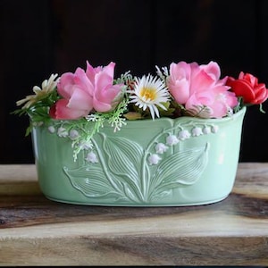 May include: A light green ceramic planter with embossed lily of the valley design, filled with pink, white, and red artificial flowers. The planter sits on a wooden surface against a dark background.