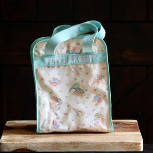 May include: A rectangular Peter Rabbit lunch bag with a light blue handle and trim. The bag features a repeating pattern of Peter Rabbit and other characters on a cream background. The text "PETER RABBIT" is embroidered on the top.