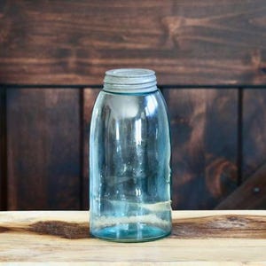 May include: A clear, vintage-style glass jar with a metal lid. The jar has a slight blue tint and is sitting on a wooden surface. The background is a dark wooden door.