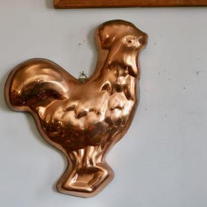 May include: A vintage copper rooster-shaped mold hanging on a white wall. The mold is made of shiny copper and has a detailed design. The rooster is a classic kitchen decor item.
