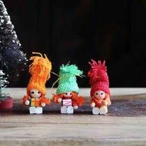 May include: Three small wooden dolls with orange, green, and red knitted hats and matching yarn hair. The dolls are standing on a wooden surface with two miniature Christmas trees in the background. The dolls are wearing colorful outfits.
