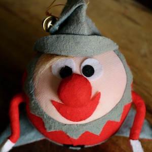 May include: A handmade Christmas elf ornament. The elf has a red nose and mouth, large black eyes, and a gray hat with a gold bell. The elf's body is red and gray, with white accents. A festive holiday decoration.