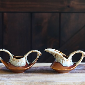 May include: Two vintage ceramic creamers with a brown and cream glaze. The creamers have a unique shape with a handle and a spout. They are displayed on a wooden surface.