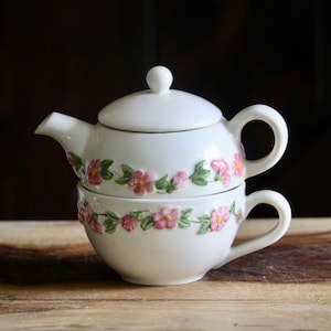 May include: A white ceramic tea-for-one set with a teapot and teacup stacked together. The set is decorated with pink floral designs and green leaves. The teapot has a lid and handle, and the teacup has a handle.