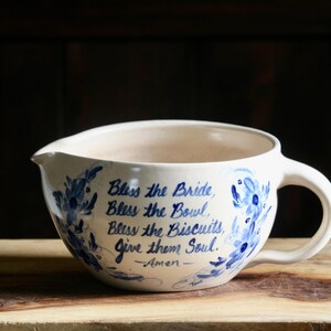 May include: A cream-colored ceramic mixing bowl with a handle and pouring spout, featuring blue hand-painted floral accents and the text: Bless the Bride, Bless the Bowl, Bless the Biscuits, Give them Soul. -Amen- written in blue script.