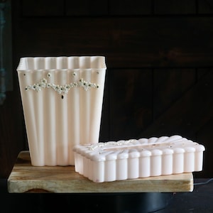 May include: A matching set of vintage, pale pink bathroom accessories. The set includes a tall, fluted wastebasket and a rectangular tissue box. Both pieces are adorned with delicate floral and rhinestone embellishments.