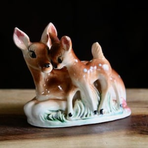 May include: A ceramic figurine of a mother deer and her fawn. The deer are brown with white spots and pink accents. They are standing on a base with green grass and a white and pink floral design. The background is dark.